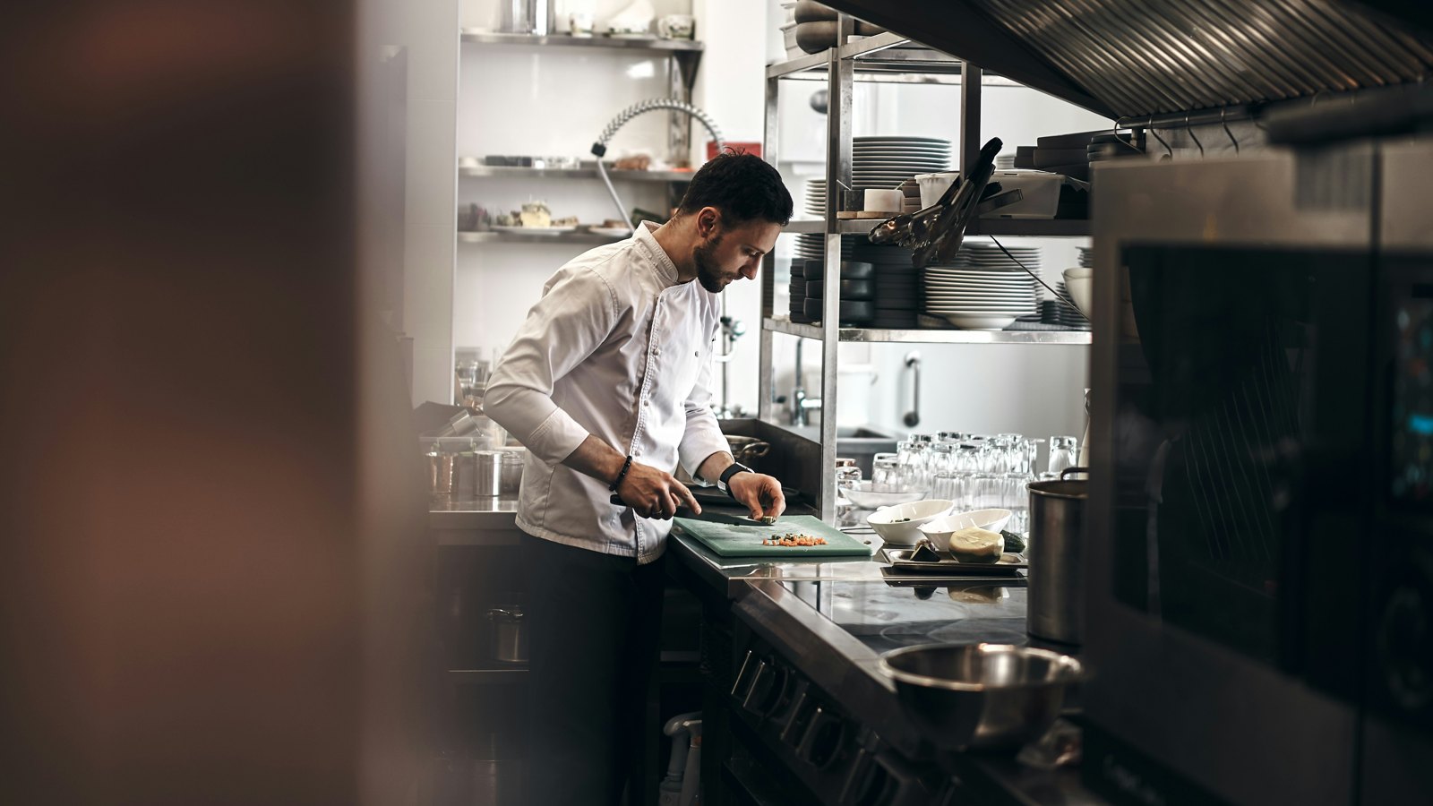 Chef preparing vegetables at a stainless steel bench beside a commercial cooking line